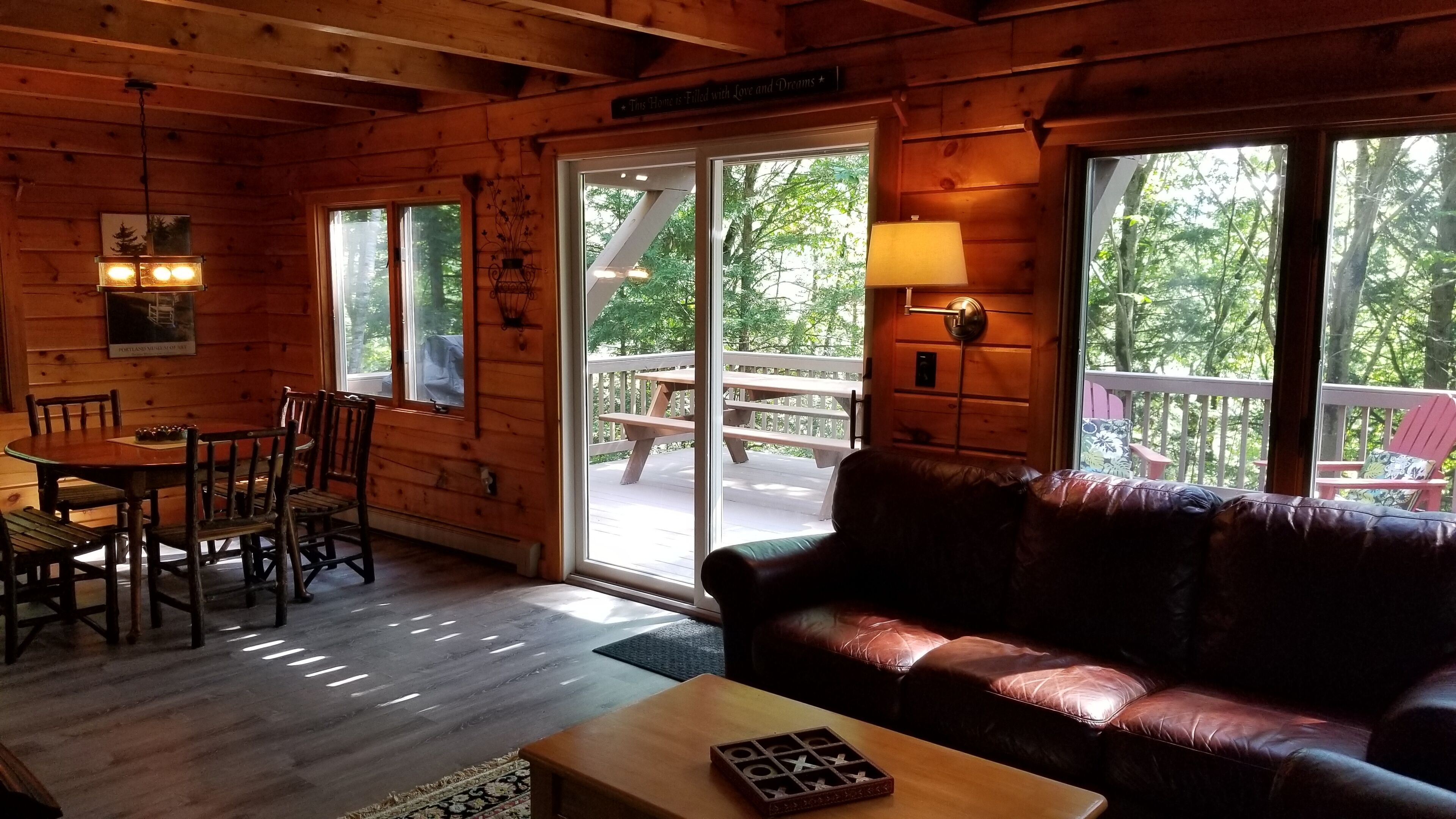 Interior acogedor de una cabaña con una mesa de comedor de madera, sillas, un sofá de cuero y un ventanal corredizo que da a una terraza exterior.