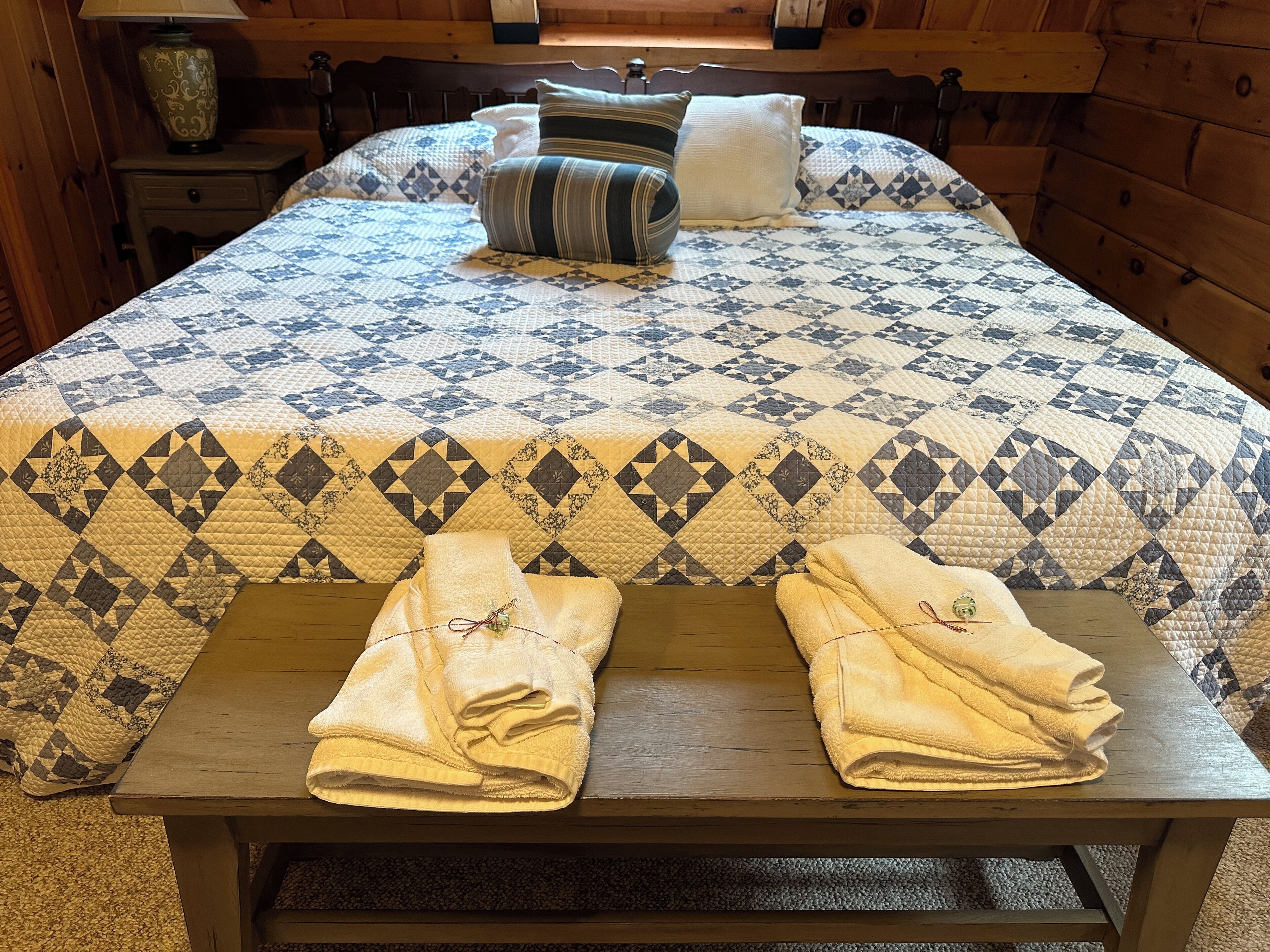A bed with a quilted bedspread, pillows, and two towels on a wooden bench.