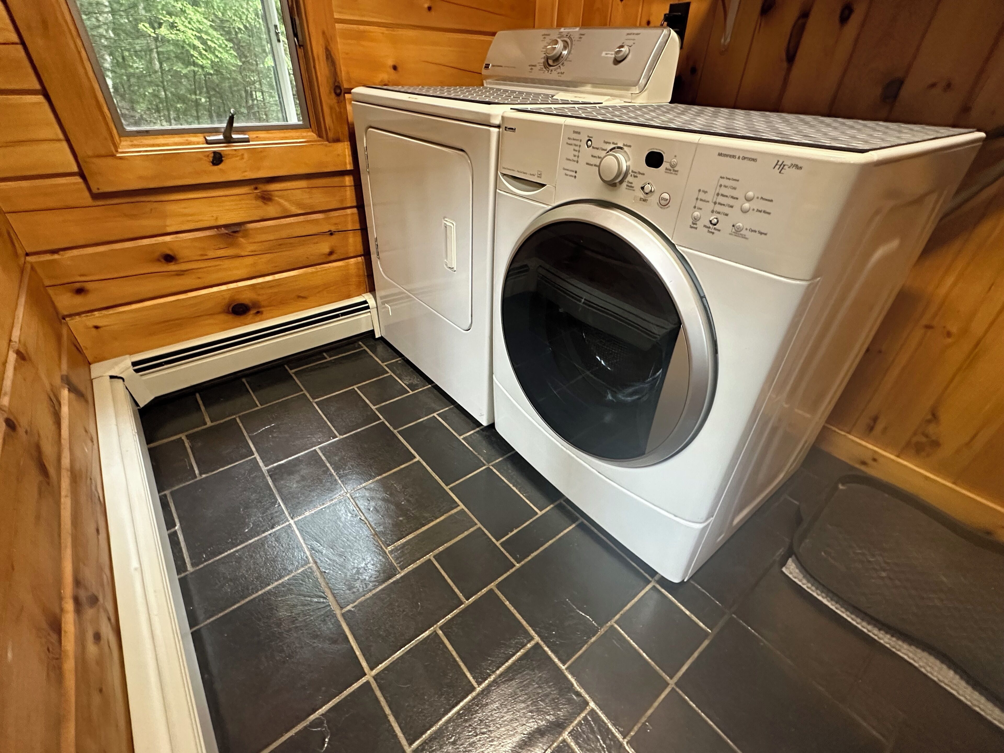 A compact laundry room with a front-loading washing machine and dryer, wooden walls, and a window with a view of trees.