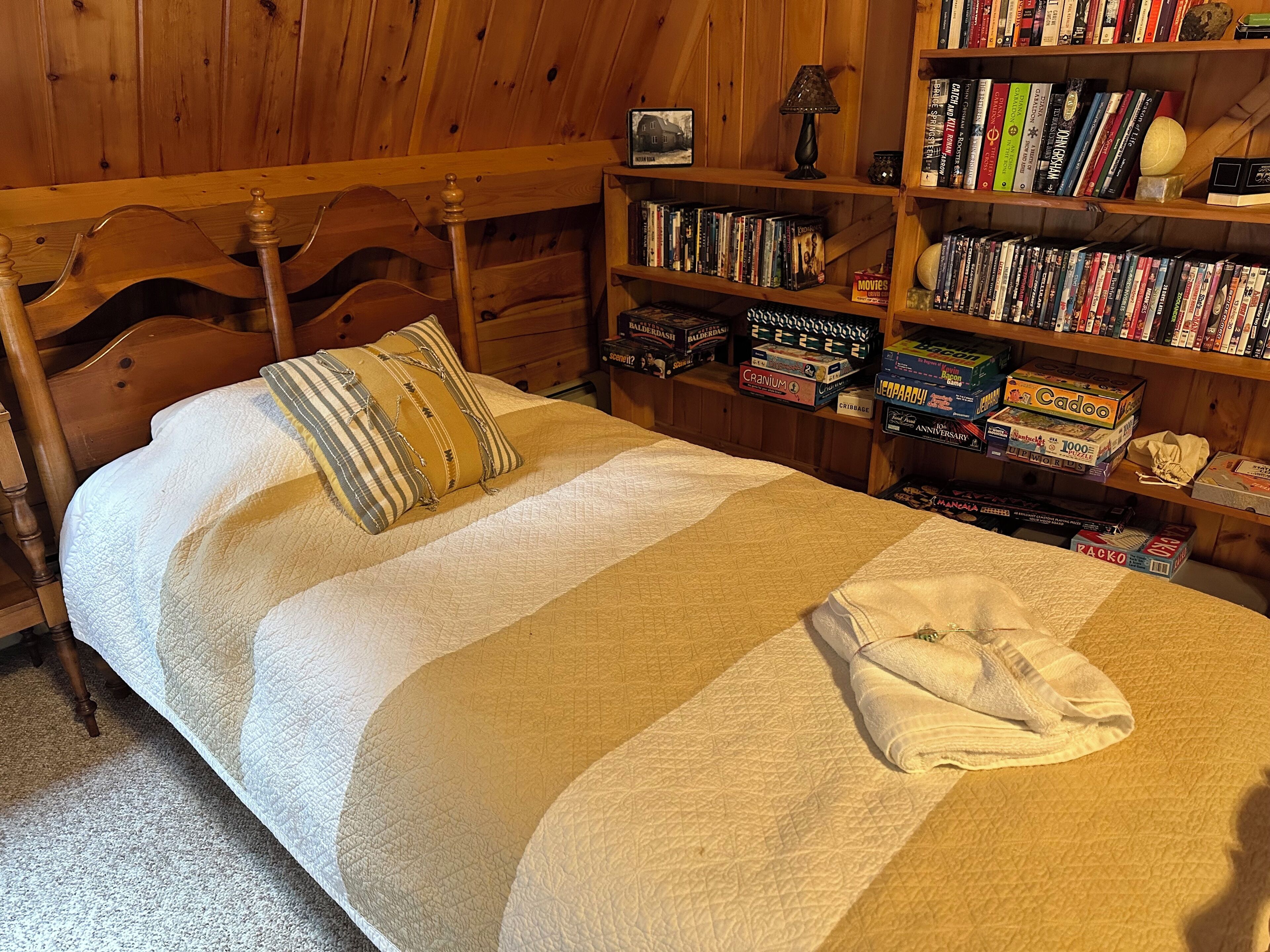 A neatly made bed with a quilt and pillows, a wooden headboard, and a bookshelf filled with books and board games.