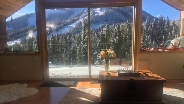 Quarto com uma grande porta de vidro deslizante que dá para uma vista nevada de montanhas. No interior, há uma mesa de madeira com um vaso de flores e um jogo de xadrez.