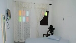 A small, simple room with a bed, a chair, and a window with curtains. - Pousada Vila do Zimbo (Cairu)