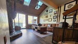 A spacious living room with a high ceiling, wooden beams, and a stone fireplace. There are multiple framed pictures on the walls and a large window providing natural light. - Cozy condo, Family- friendly just completely remodeled. SKI IN -SKI OUT!! (Champion)