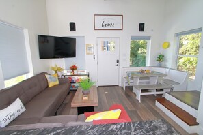 A compact living space with a sofa, a coffee table, a TV, a dining table with benches, and a door with a window. - Hot Tub!•FirePit-Table•King-Bed•Walk to "The Island"•Trailer-Parking! (Pigeon Forge)