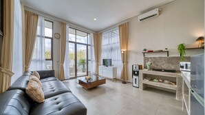 A modern living room with a sofa, a coffee table, and a TV. There is a kitchen area with a fridge, microwave, and a plant. - Residence at Damara Village Jimbaran (Jimbaran)