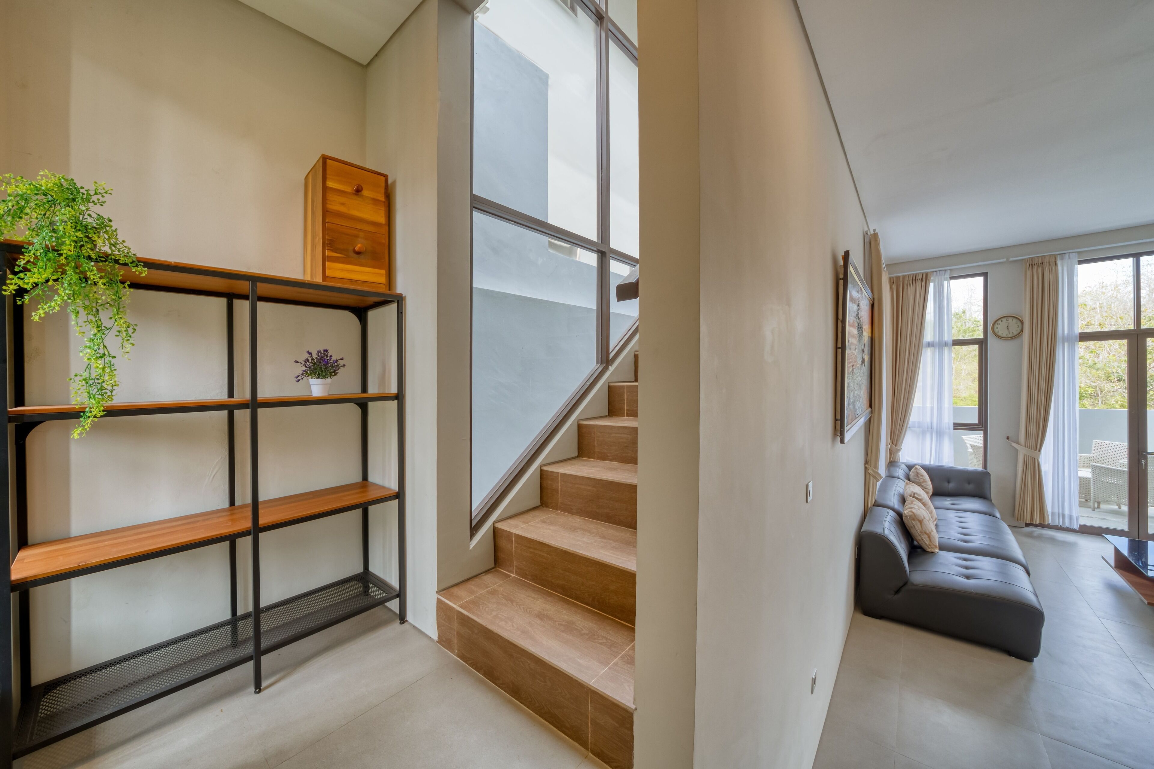 A modern interior with a staircase, a wooden shelf with plants, a grey sofa, and a large window.