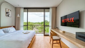 A hotel room with a bed, a desk, a chair, a TV mounted on the wall, and a large window with a view of greenery. - Residence at Damara Village Jimbaran (Jimbaran)