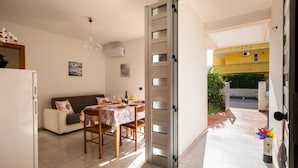 Puerta abierta que da a un balcón con vistas a un edificio amarillo, zona de comedor con una mesa preparada para dos, un sofá y un refrigerador.