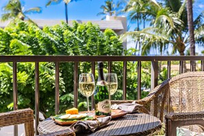 A wicker table with a bottle and glasses of white wine, a plate with cheese, and a napkin on a balcony with a view of palm trees and a building. - Kihei Akahi D205 | Steps to Kamaole II Beach (Kihei)