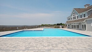 A rectangular swimming pool with a paved deck, surrounded by a wooden fence, and a large house with a gabled roof and multiple windows. - 2507 - Salty Sandpiper - Coastal Retreat in Duck | Hot Tub & Sunsets (Duck)