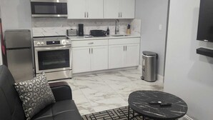 A modern kitchen with white cabinets, a microwave, stove, and a sink. There's a black sofa with a patterned pillow, a small round table, and a trash can. - Majestically Pleasant Suite (Hempstead)