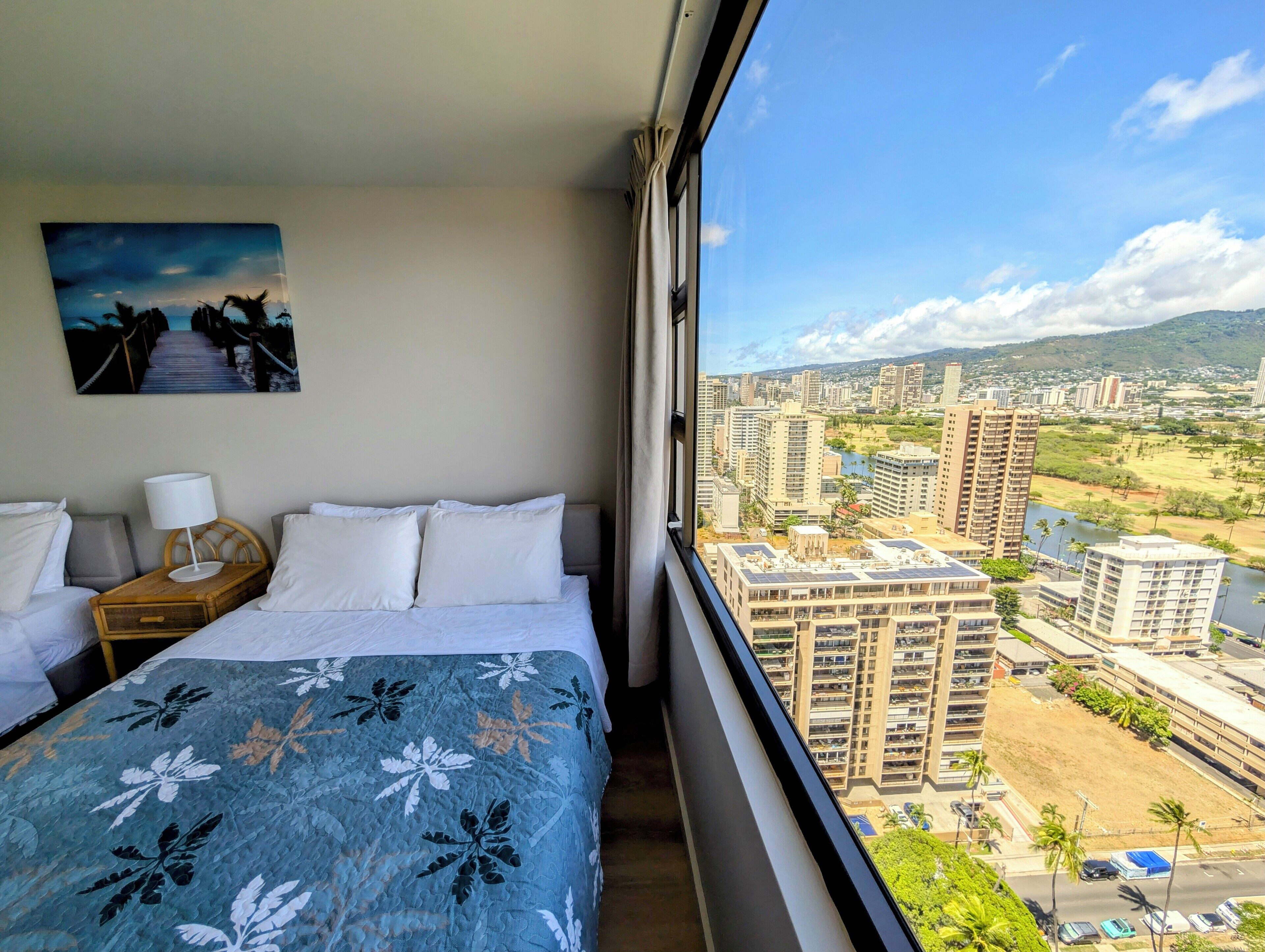 Hotelkamer met een bed, nachtkastje, lamp en schilderij. De kamer heeft een groot raam met uitzicht op een stadsgezicht met gebouwen en een rivier.