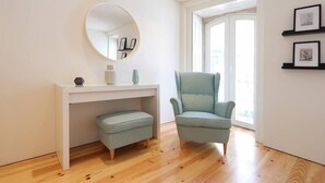 A modern room with a white desk, a round mirror, a light blue armchair, and a small ottoman. There are framed pictures on the wall and a large window providing natural light. - HM - Pinheiro Duplex Apartment (Porto)
