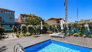 Une piscine dotée d’une terrasse en bois, entourée de verdure et de bâtiments.