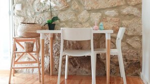 Une pièce avec un mur en pierre, une table blanche, deux chaises blanches, une chaise en bois, un panier en osier, une plante en pot et un petit vase rose.