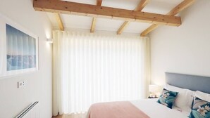 Ein Schlafzimmer mit einem Bett, einem Fenster mit Vorhängen, einem Heizkörper und einem Holzbalken an der Decke.