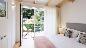 Ein Schlafzimmer mit Bett, Nachttisch, Lampe, einem Fenster mit Vorhängen und Blick auf einen Garten.