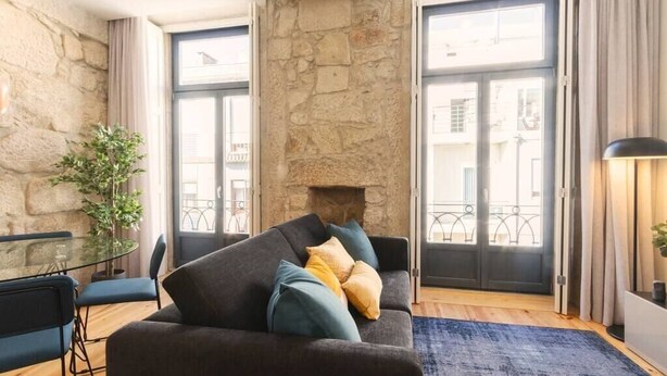 A living room with a stone wall, a sofa with cushions, a glass table with chairs, and a view through a glass door. - HM – Oporto Downtown Swimming Pool Apartment (Porto)