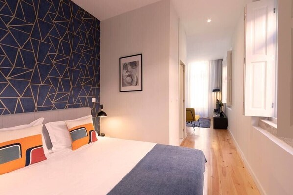A hotel room with a geometric-patterned headboard, a bed with colorful pillows, a framed artwork on the wall, and a hallway leading to another room.
