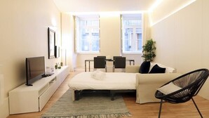 A modern hotel room with a bed, a TV on a white console, a dining table with chairs, and a window with a view of buildings. - HM - Ribeira São João Apartment - 2D (Porto)
