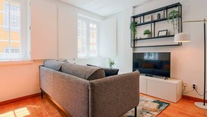 Una sala de estar moderna con un sofá gris, un televisor de pantalla plana sobre un mueble blanco, una estantería negra con plantas y una lámpara de pie.