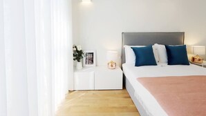Een moderne slaapkamer met een groot bed, witte nachtkastjes en een nachttafeltje met een lamp.