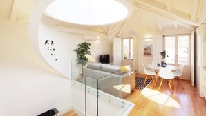 A modern living room with a glass staircase, a grey sofa, a dining area with a white table and chairs, and a large window providing natural light.