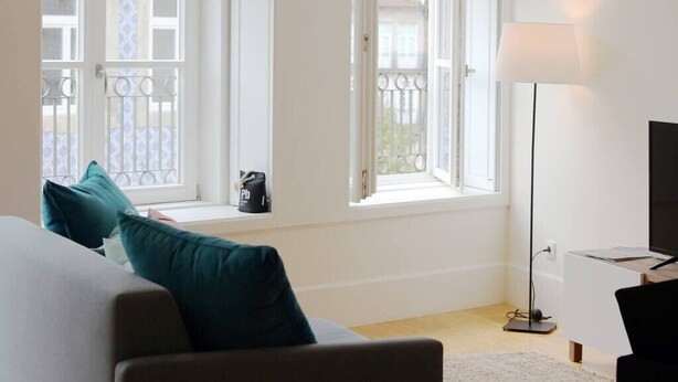 A room with a sofa, a standing lamp, and a TV. There are windows with white frames and a balcony visible through the windows. - HM - Carlos Alberto Apartment 2 (Porto)