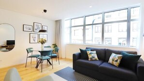 A modern living room with a dark sofa, teal chairs, a dining table, and a large window with a city view. - HM - Sta Catarina Charming Apartment (Porto)