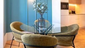 Ein Essbereich mit einem Glastisch und zwei Stühlen, einer Vase mit Blumen sowie eine Küche im Hintergrund.