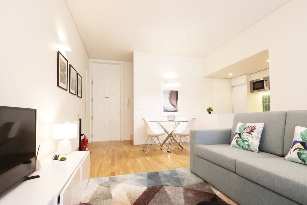 A modern living room with a flat-screen TV, a grey sofa, and a dining area with a glass table and chairs.