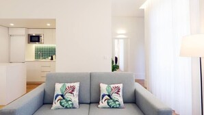 A modern living room with a grey sofa, two decorative pillows, a flat-screen TV, and a kitchen in the background.