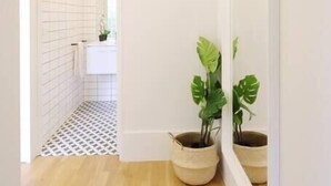 A hallway with a wooden floor, white walls, and a white tiled area leading to a bathroom. There are two potted plants on the floor.