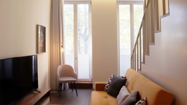 Una sala de estar con una televisión de pantalla plana, un sofá con cojines, una silla y una escalera.
