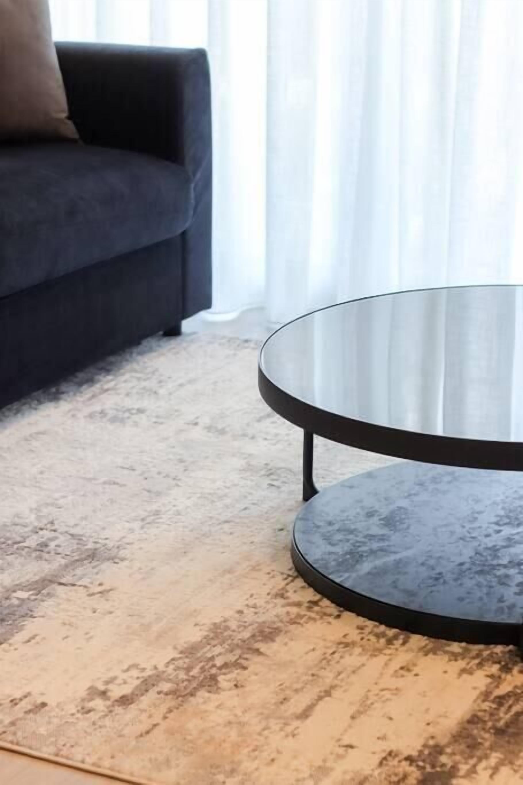 Une table basse ronde en verre, dotée d’un cadre en métal noir, placée sur un tapis beige à texture.