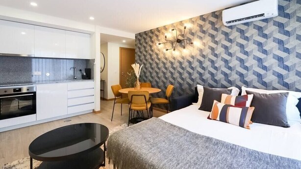 Une chambre d’hôtel moderne avec un lit, une table de chevet et un petit pouf noir. On y trouve une kitchenette équipée d’une cuisinière et d’un four. Un coin repas avec une table et des chaises est visible au fond.