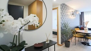 Une chambre d’hôtel moderne avec un miroir rond, des orchidées blanches, une petite table avec une assiette et une bougie, un coin repas avec des chaises jaunes et un mur à motifs.