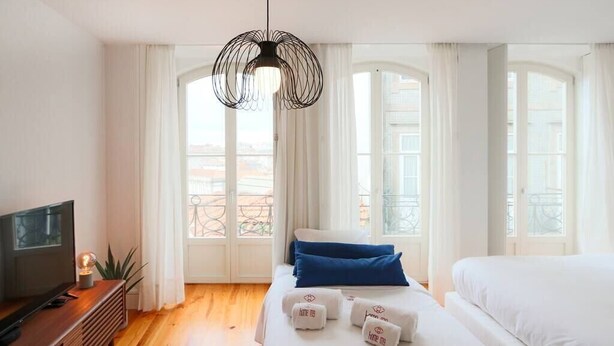 A hotel room with a bed, a TV, and a view of the city through large windows. - HM – Historic Downtown Batalha Studio (Porto)