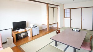 Ein traditioneller japanischer Raum mit Tatami-Böden, einem niedrigen Tisch, einem Fernseher, einem kleinen Kühlschrank und Schiebetüren.