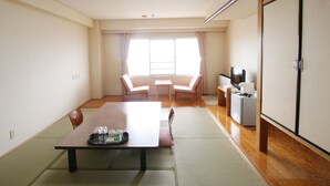 Ein Hotelzimmer mit Tatami-Boden, einem Holztisch mit Tablett, Gläsern und einer Flasche, zwei Stühlen, einem Fernseher und einem Schrank.
