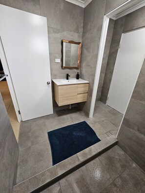 A bathroom with a white sink, wooden vanity, a mirror, and a blue rug. - Yiayi's House Getaway (Wagaman)