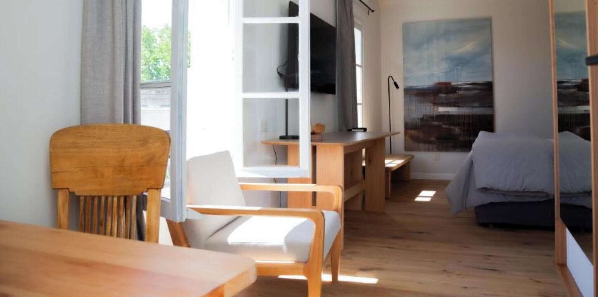 A room with a wooden desk, chairs, a bed, a TV, and a painting on the wall.