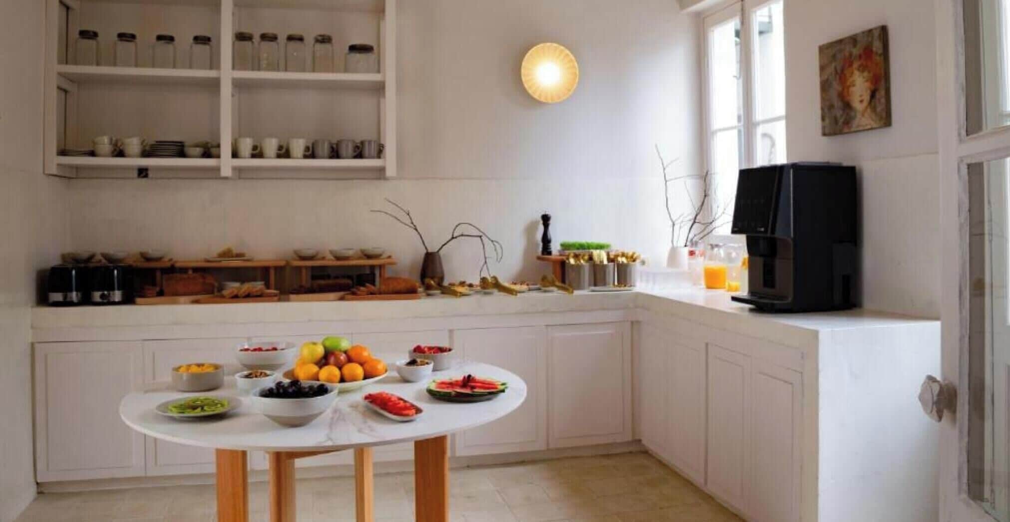 A kitchen with white cabinets, a round dining table with fruit, a coffee maker, and a window with a painting.