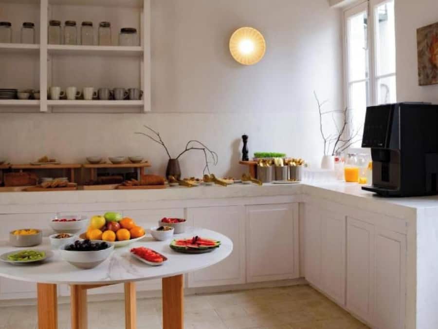 A kitchen with white cabinets, a round dining table with fruit, a coffee maker, and a window with a painting.