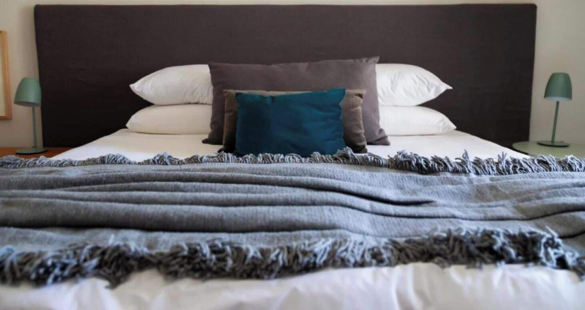 Een bed met een donker hoofdbord, witte kussens, een blauw accentkussen en een grijze deken.