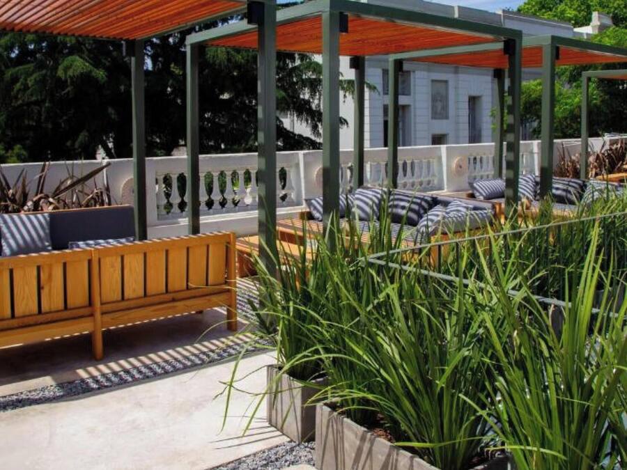 A covered outdoor seating area with a wooden bench and lounge chairs, surrounded by tall plants and a pool.