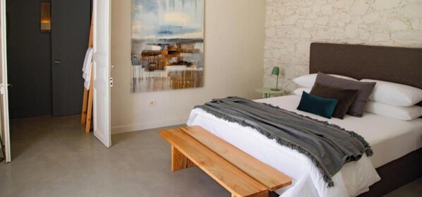 Um quarto com cama, um banco, um quadro, um abajur e uma parede de pedra.
