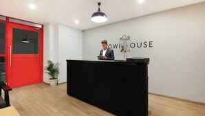 Un comptoir d’accueil avec une personne derrière, une porte rouge, une plante en pot et un mur sur lequel on peut lire le texte « OWI HOUSE ».