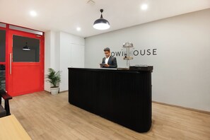 Een receptiebalie met een persoon erachter, een rode deur, een potplant en een muur met de tekst 'OWI HOUSE'.