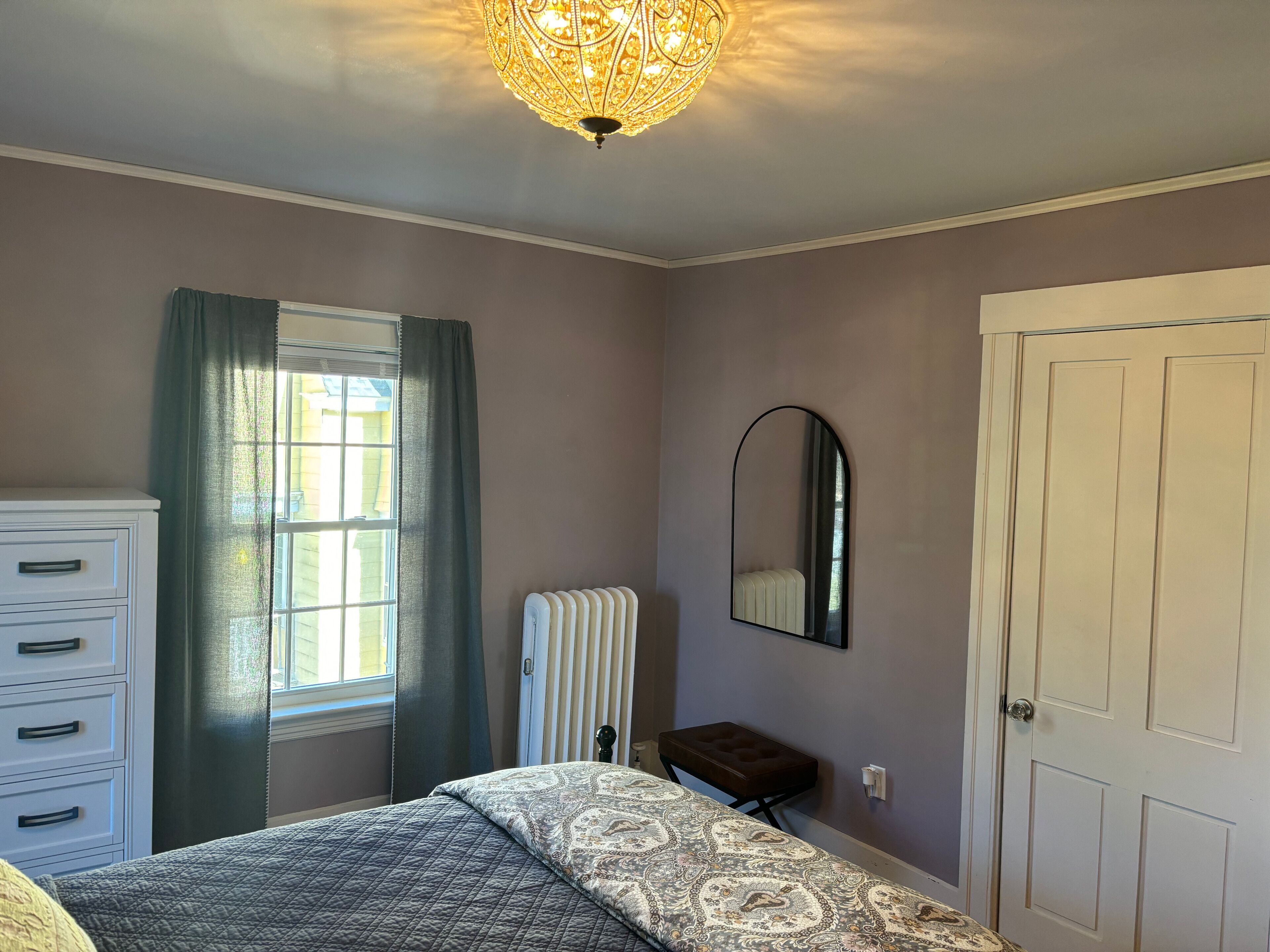 A bedroom with a bed, a dresser, a radiator, a mirror, and a door.